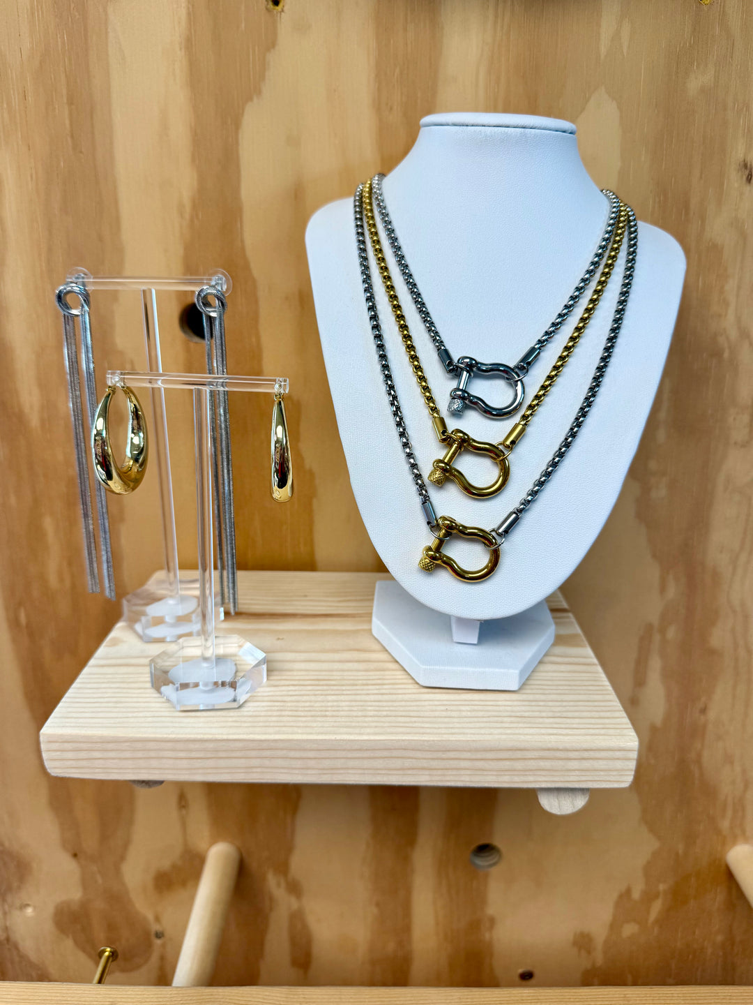 Three necklaces on a stand, each with a chunky square-link chain and a central snaffle bit clasp, in mixed metal, gold, and silver finishes.